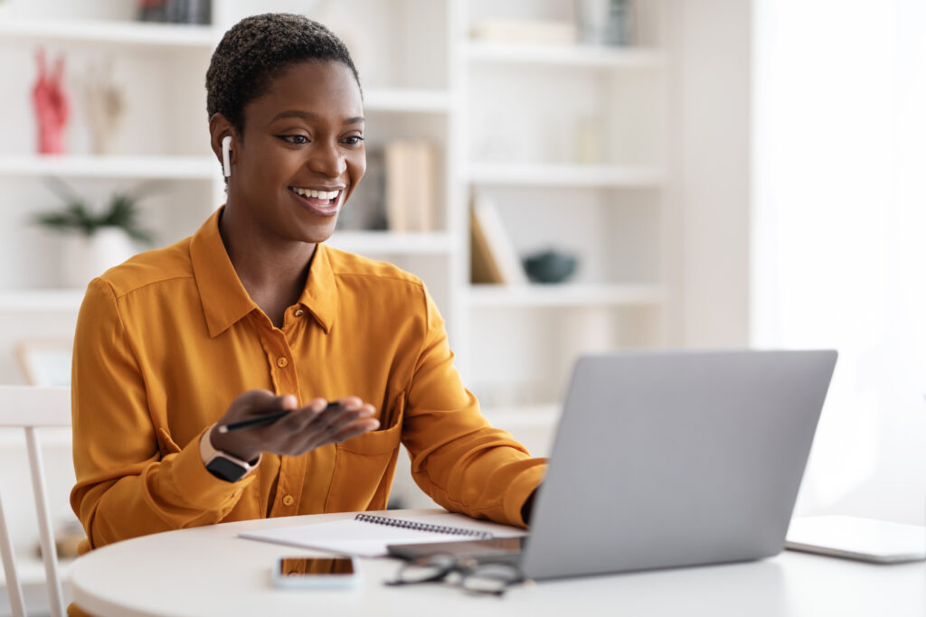 Attractive young African American woman having video conference with clients or business partners, sitting at work desk at office, using laptop and earbuds, gesturing and smiling.