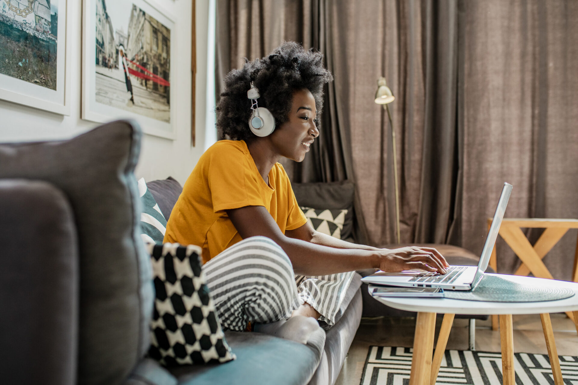 A young African American woman with wireless headphones is using a laptop and working in the living room while smiling and discussing the work on a video.