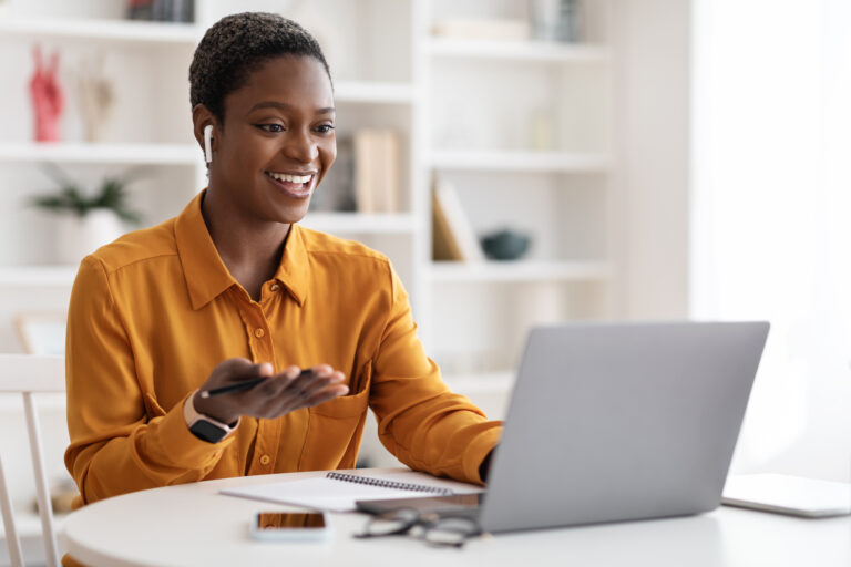Attractive young African American woman having video conference with clients or business partners, sitting at work desk at office, using laptop and earbuds, gesturing and smiling.