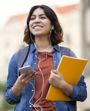 Cheerful young arab student female listening music with smartphone and earphones outdoors, smiling middle eastern woman carrying workbooks and backpack while walking on city street.