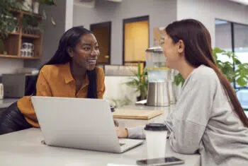 Two Young Businesswomen Working On Laptop In Kitchen Area Of Modern Office