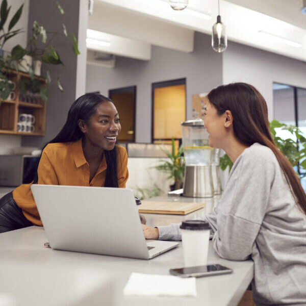 Two Young Businesswomen Working On Laptop In Kitchen Area Of Modern Office Two Young Businesswomen Working On Laptop In Kitchen Area Of Modern Office