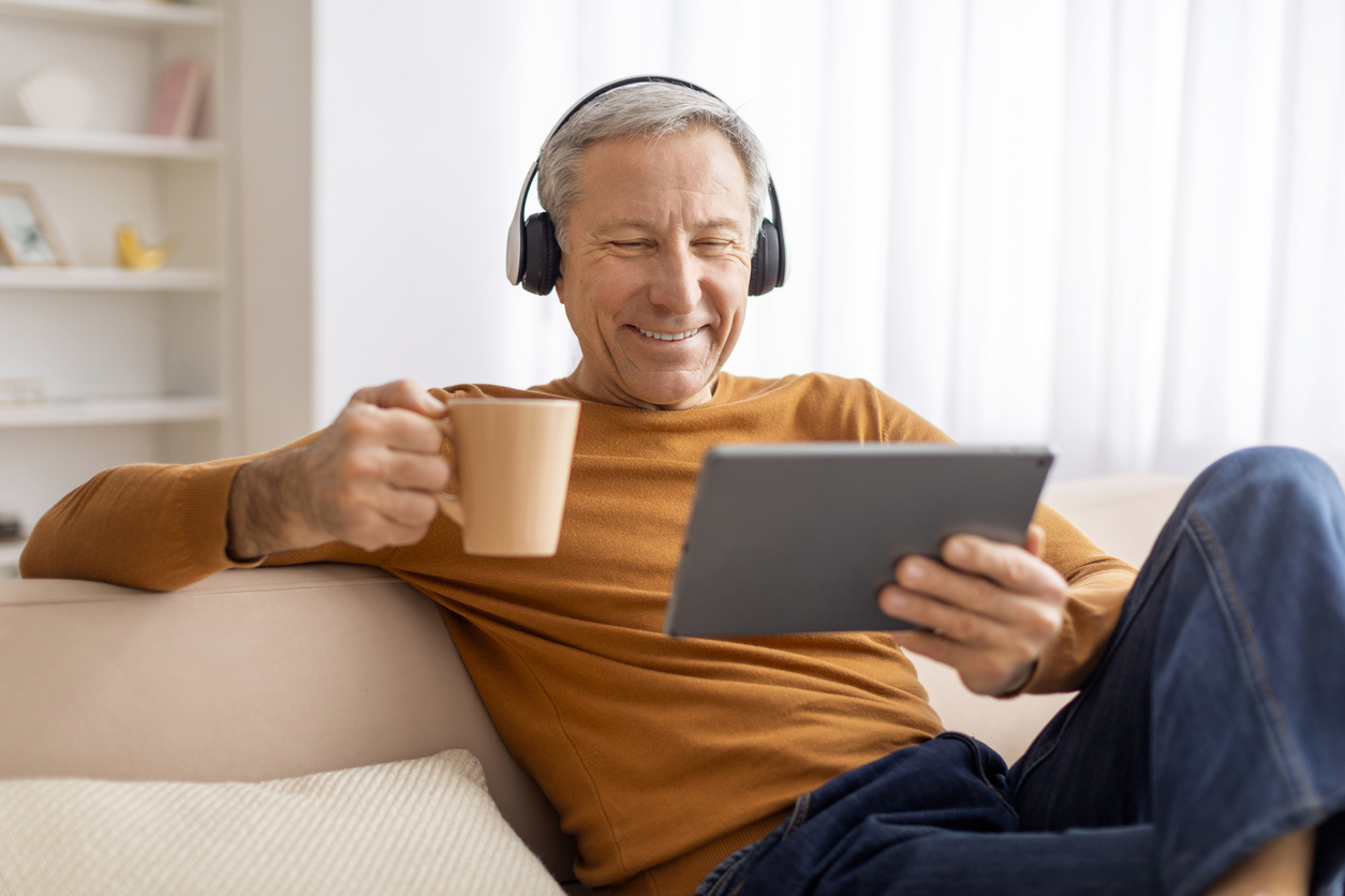 Relaxed elderly man chilling at home at weekend, using digital tablet and wireless headphones, watching movie online and drinking coffee, sitting on couch at home, copy space