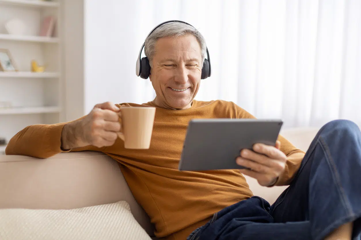 Relaxed elderly man chilling at home at weekend, using digital tablet and wireless headphones, watching movie online and drinking coffee, sitting on couch at home, copy space