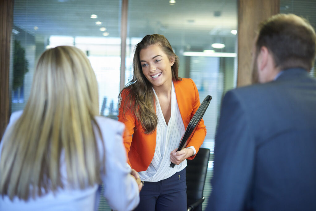 A young graduate walks into an interview room full of confidence and positivity energy. She is holding her cv and smiling at the interview panel before her. She is wearing blue trousers with an orange suit jacket , as she opens the door and strides in and shakes hands with a woman on the panel. in the foreground we can see the back of two of the panel, a man and a woman.