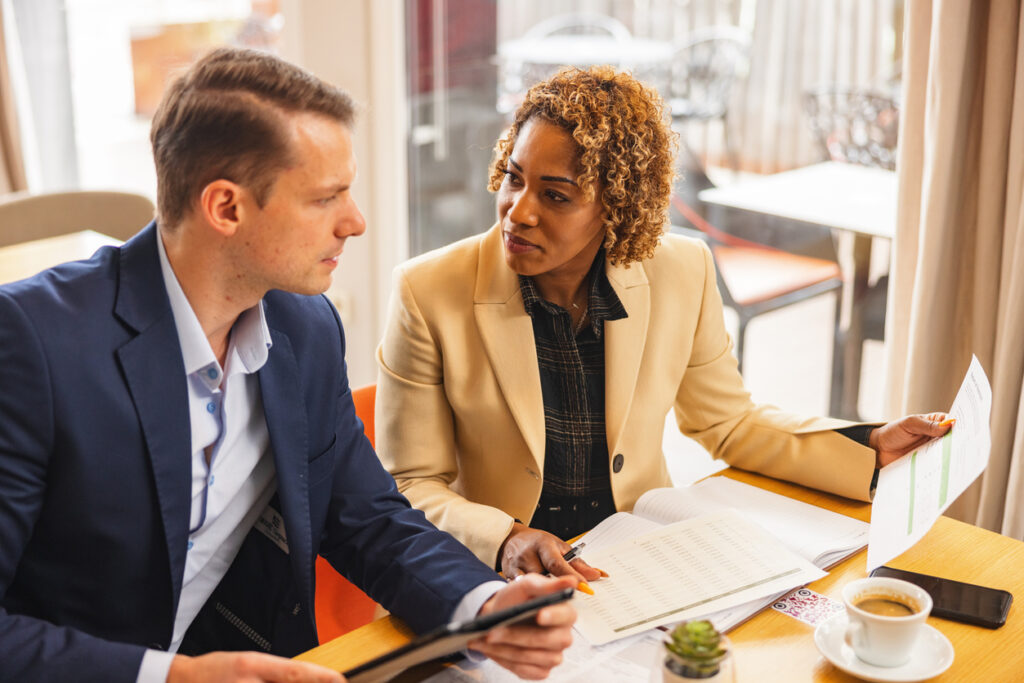 A Caucasian Businessman And A Black Businesswoman Going Over Annual Reports While At The Corporate Cafeteria A professional mid-adult businessman looking at the documents together with his female colleague. They are sitting in a corporate cafeteria and discussing the numbers and strategies. They look serious and focused. They are wearing businesswear.