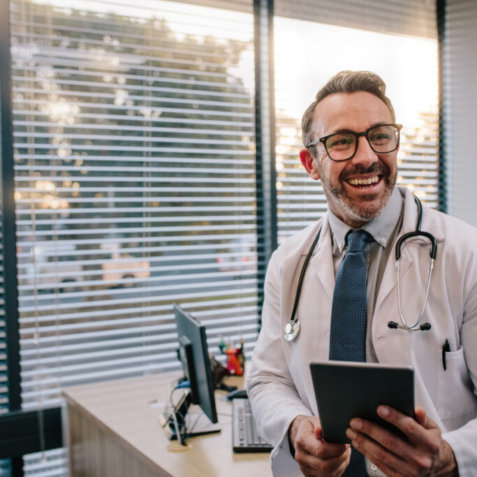 Doctor with digital tablet in his office Smiling mature male doctor with digital tablet in his office. Friendly medical professional with tablet computer in clinic.
