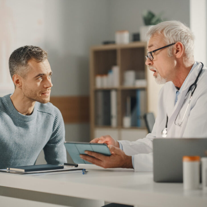 Experienced Middle Aged Family Doctor Showing Analysis Results on Tablet Computer to Male Patient During Consultation in a Health Clinic. Physician Sitting Behind a Desk in Hospital Office. Experienced Middle Aged Family Doctor Showing Analysis Results on Tablet Computer to Male Patient During Consultation in a Health Clinic. Physician Sitting Behind a Desk in Hospital Office.