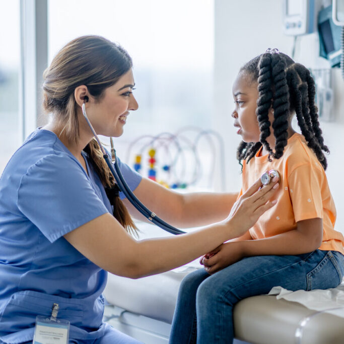 Children's Medical Appointment A sweet little girl of African decent, sits up on an exam table during a routine check-up. She is dressed casually and is smiling gently as her doctor leans in with her stethoscope to listen to her heart.